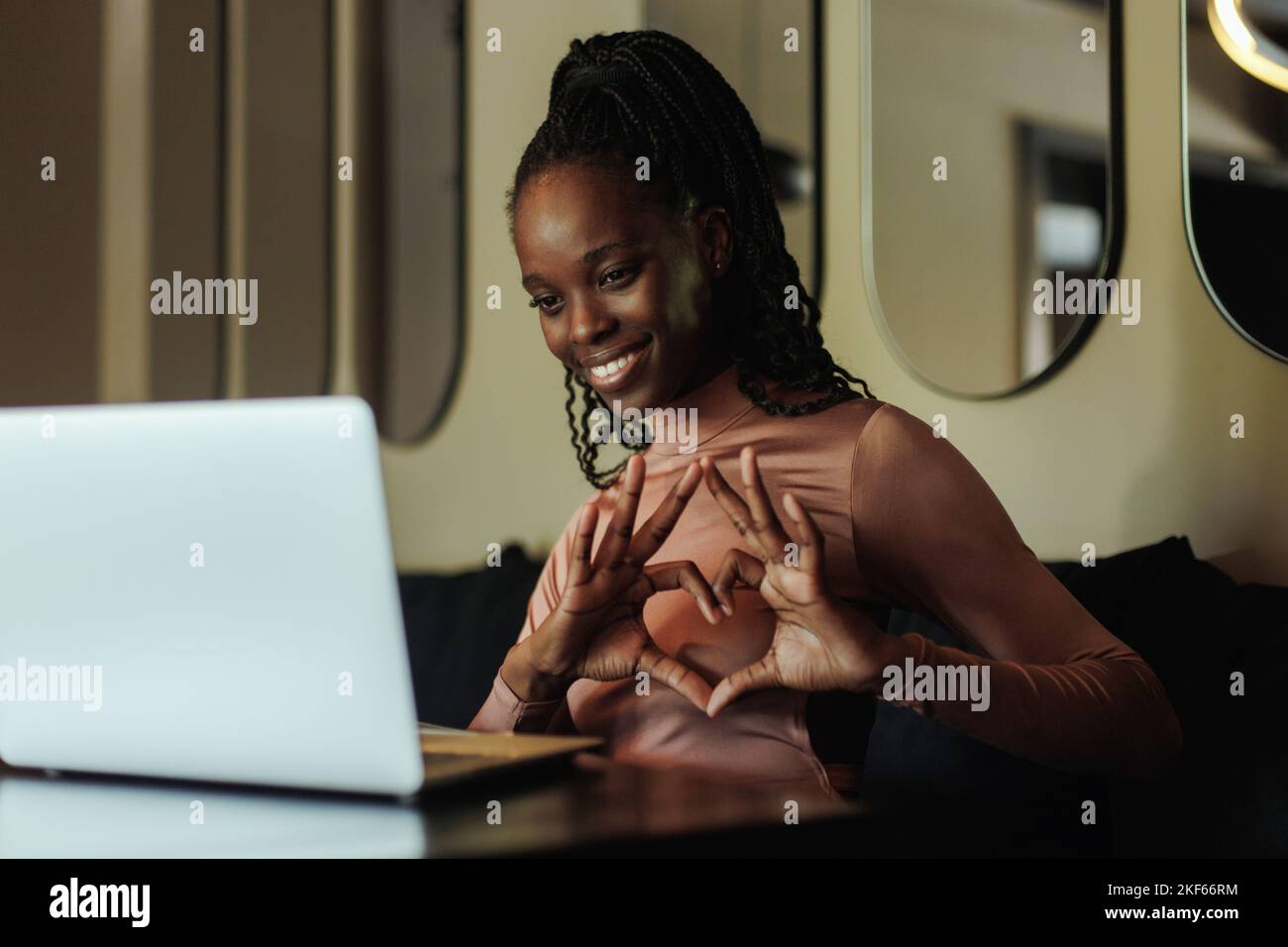 Ritratto di una giovane donna afroamericana sorridente seduta al tavolo vicino al computer portatile, che mostra il cuore con le dita sullo schermo. Foto Stock