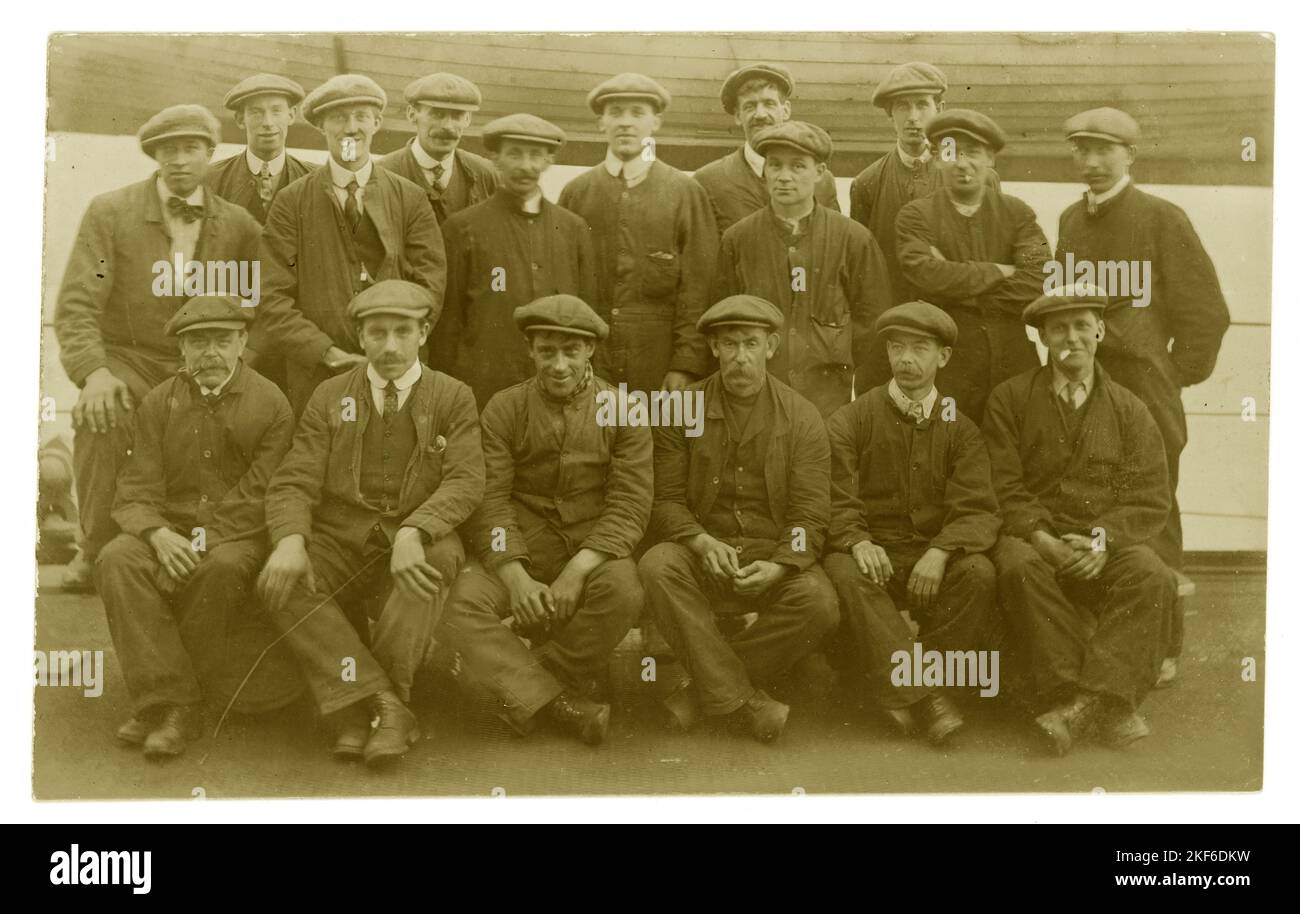 Originale post del 1900 WW1 cartolina di uomini della classe operaia, operai, indossando le tipiche calotte piatte, tute, del periodo, circa 1919 e più tardi. Forse lavoratori portuali, costruttori navali, portieri, come scafo di legno in background, molti personaggi, Regno Unito Foto Stock