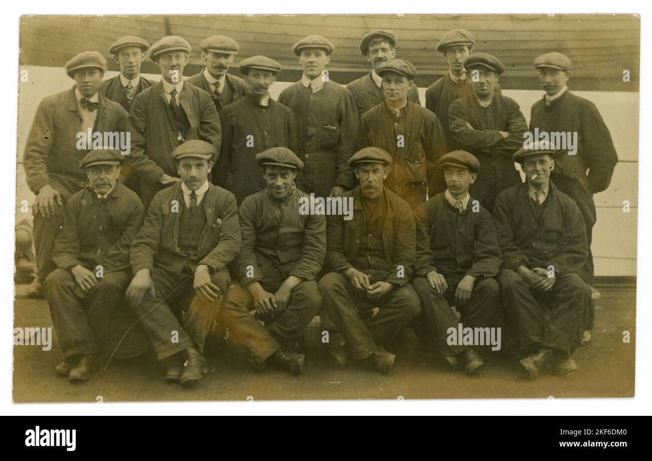 Originale post del 1900 WW1 cartolina di uomini della classe operaia, operai, indossando le tipiche calotte piatte, tute, del periodo, circa 1919 e più tardi. Forse lavoratori portuali, costruttori navali, portieri, come scafo di legno in background, molti personaggi, Regno Unito Foto Stock