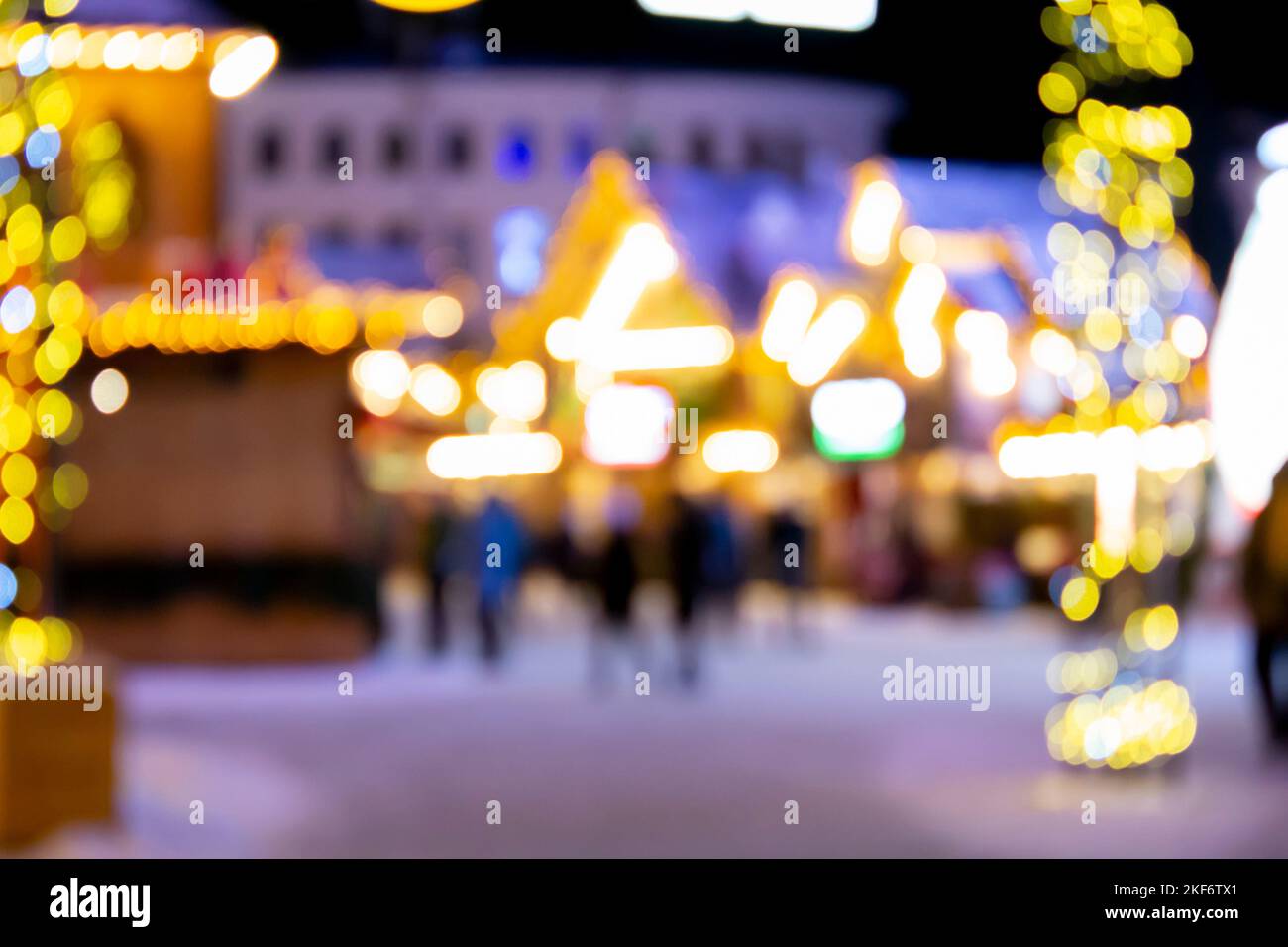 Sfondo sfocato. La gente cammina nella piazza della città la sera d'inverno. Silhouette nere di persone che camminano vicino a case decorate illuminazione luminosa. Luce bianca bokeh sfocare i punti dalle luci della casa incandescente Foto Stock