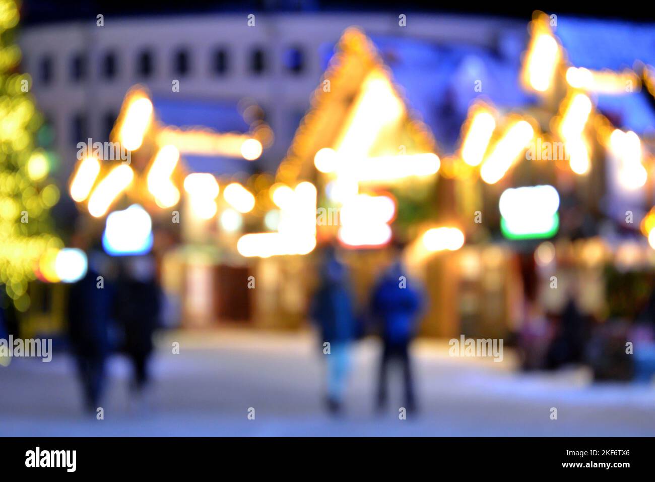 Sfondo sfocato. La gente cammina nella piazza della città la sera d'inverno. Silhouette nere di persone che camminano vicino a case decorate illuminazione luminosa. Luce bianca bokeh sfocare i punti dalle luci della casa incandescente Foto Stock