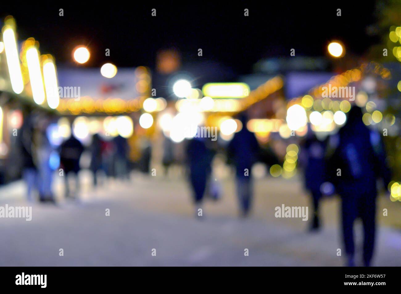 Sfondo sfocato. La gente cammina nella piazza della città la sera d'inverno. Silhouette nere di persone che camminano vicino a case decorate illuminazione luminosa. Luce bianca bokeh sfocare i punti dalle luci della casa incandescente Foto Stock