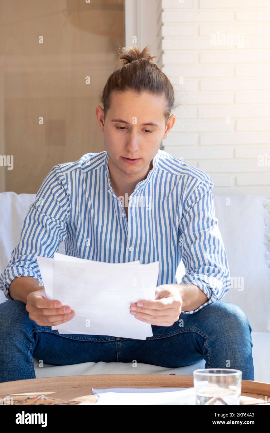Vista verticale del giovane uomo con un'acconciatura da mordente che indossa una camicia bianca e blu a righe che legge importanti documenti contrattuali nel soggiorno Foto Stock
