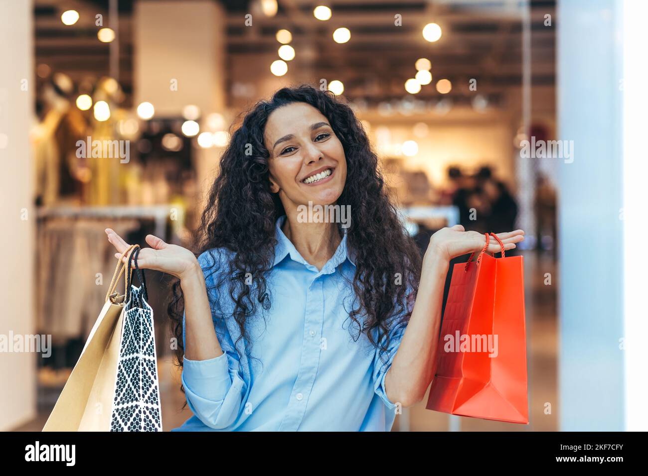 Una giovane donna felice spagnola, latino-americana sta facendo shopping di festa in un supermercato, comprando i regali per lei e la sua famiglia per le feste di Natale. Tiene sacchetti di carta colorati con le merci. Foto Stock