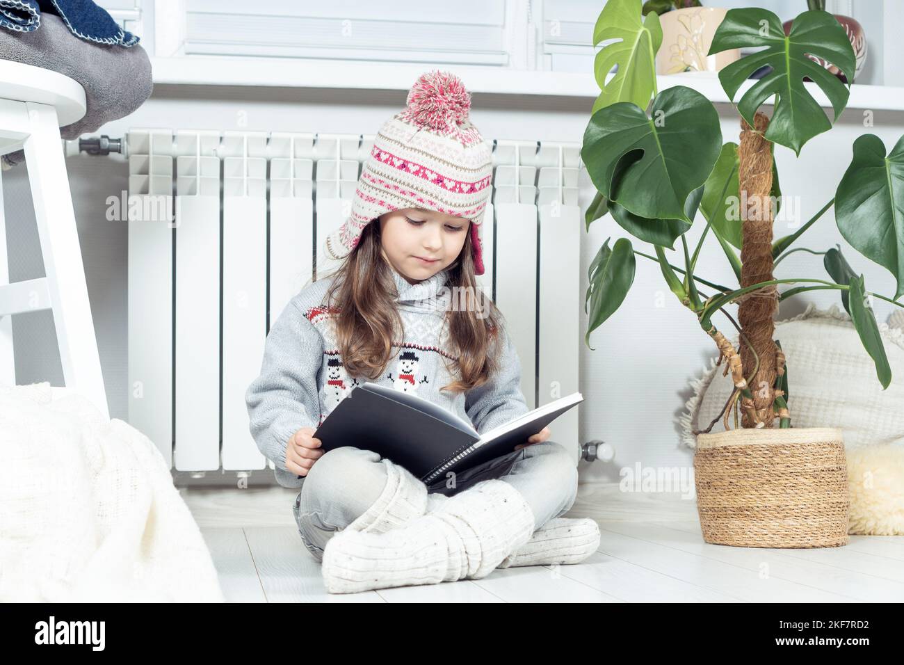 Interessato, ragazza carina seria in cappello di lana e vestiti caldi con libro di lettura del pupazzo di neve vicino al radiatore di riscaldamento in soggiorno bianco. Accogliente e Foto Stock