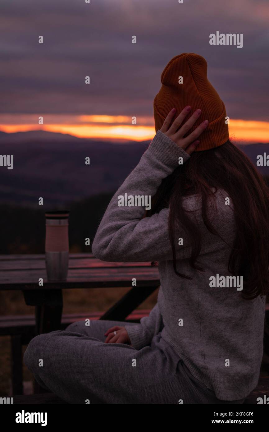 La giovane donna è seduta con thermos alla panchina e guarda la vista delle montagne al tramonto Foto Stock