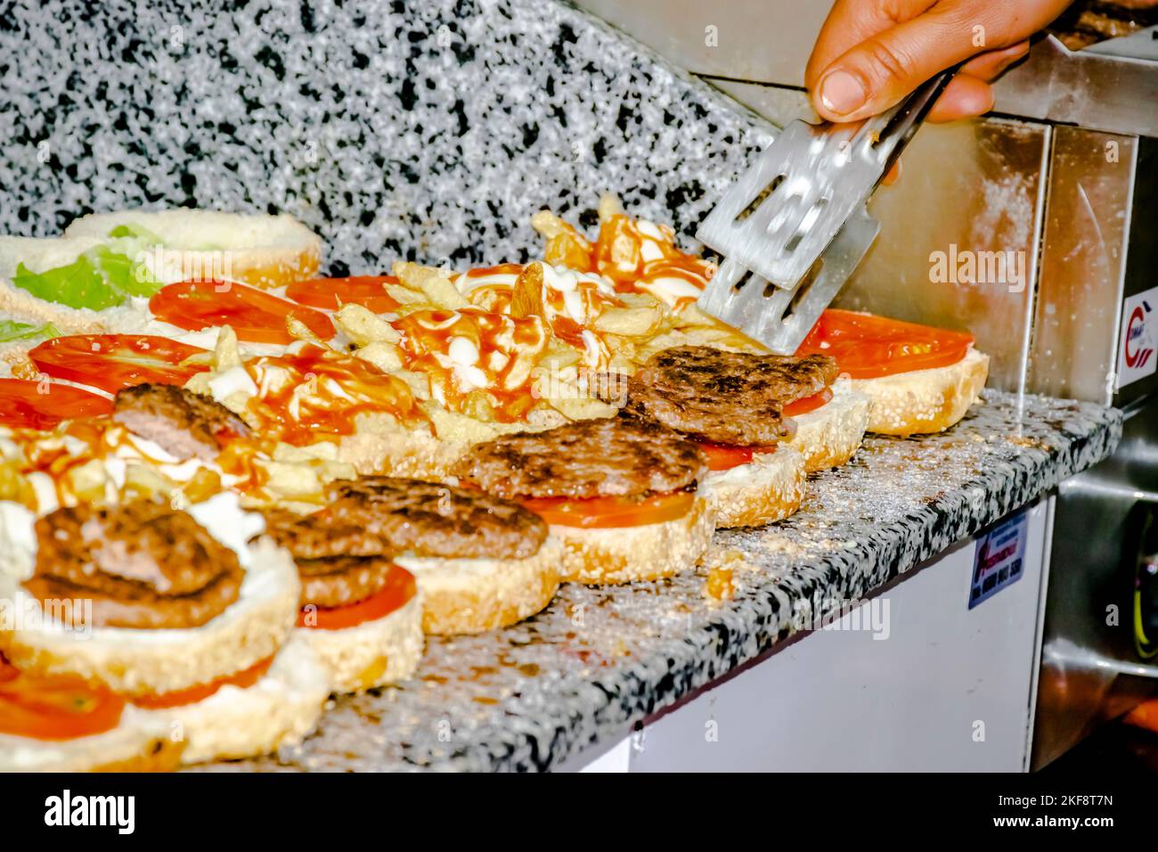 Focalizzazione selettiva sulla mano del cuoco, sulle tenaglie e sugli hamburger ripieni di patatine fritte, lattuga, pomodoro, bistecca alla griglia tritata, maionese e salsa ketchup. Foto Stock