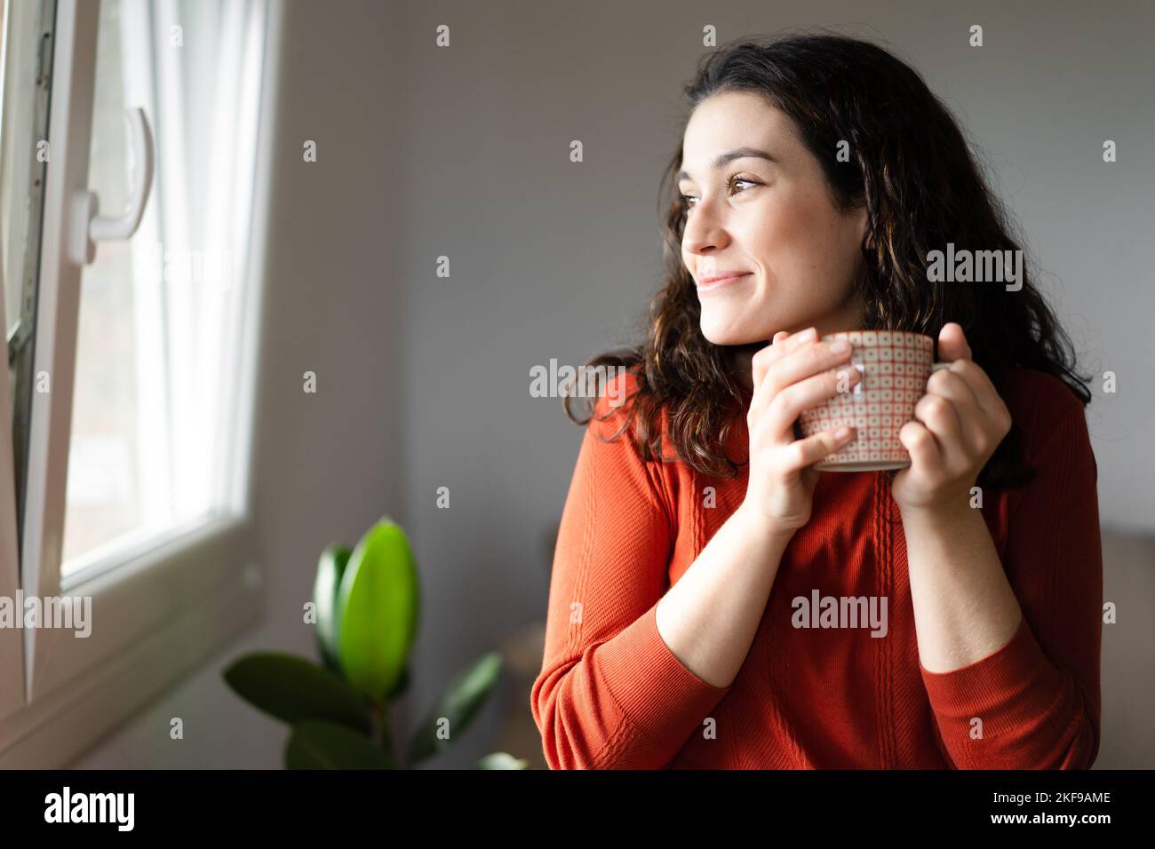 Giovane donna felice bella tenendo una tazza di bevanda calda e guardando attraverso la finestra sentirsi bene Foto Stock