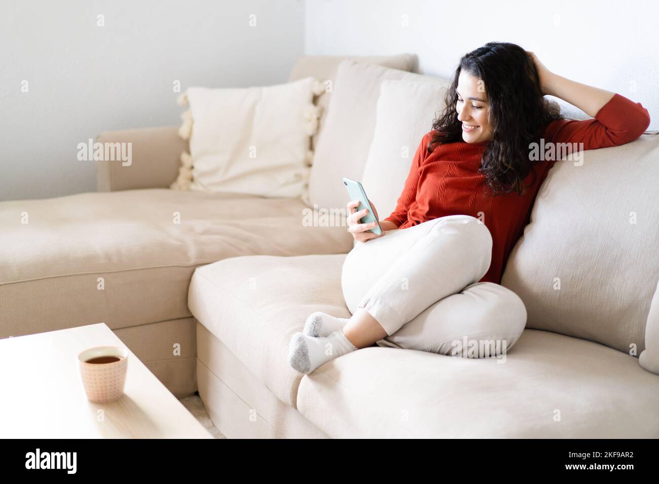 Giovane donna felice seduta comodamente sul divano utilizzando il suo smartphone e bevendo una bevanda Foto Stock