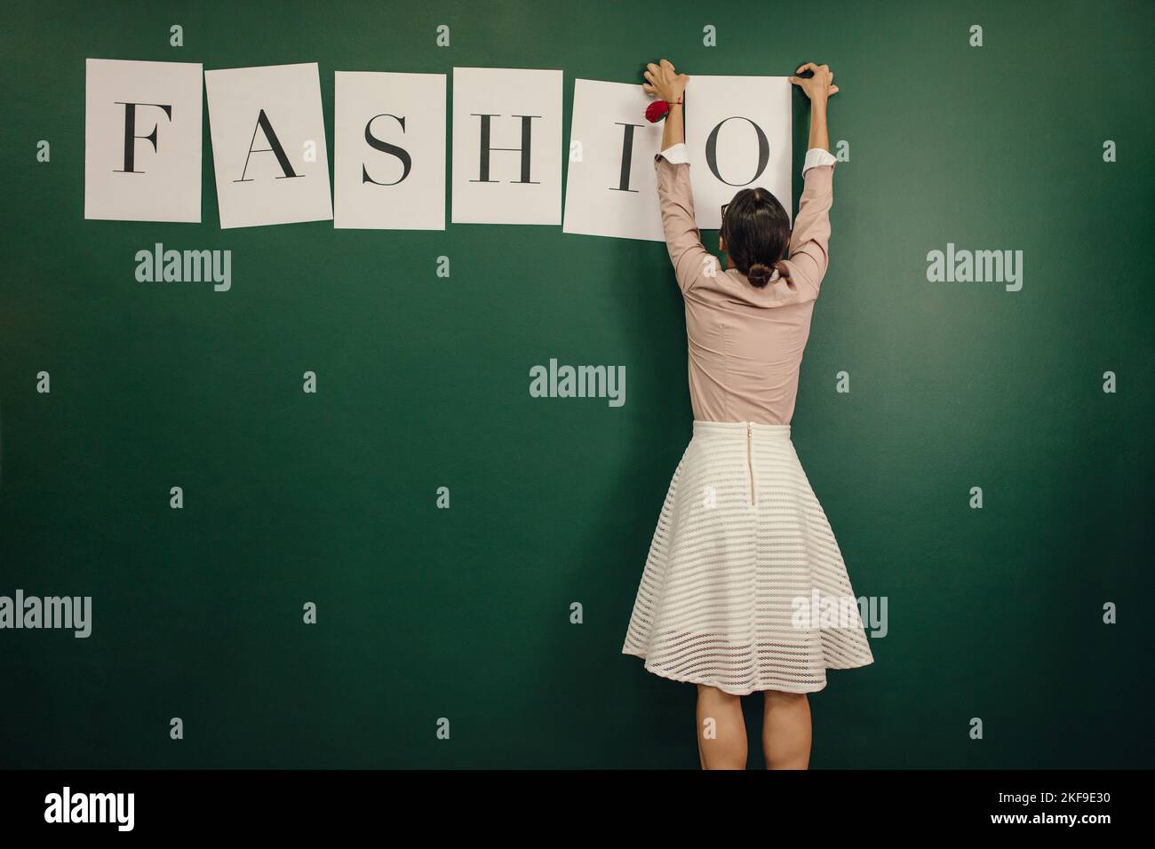 Vista posteriore ripresa di una giovane donna che organizza lettere di parole moda sulla parete. Una designer femminile che si appiccicava la parola sulla parete del suo studio. Foto Stock