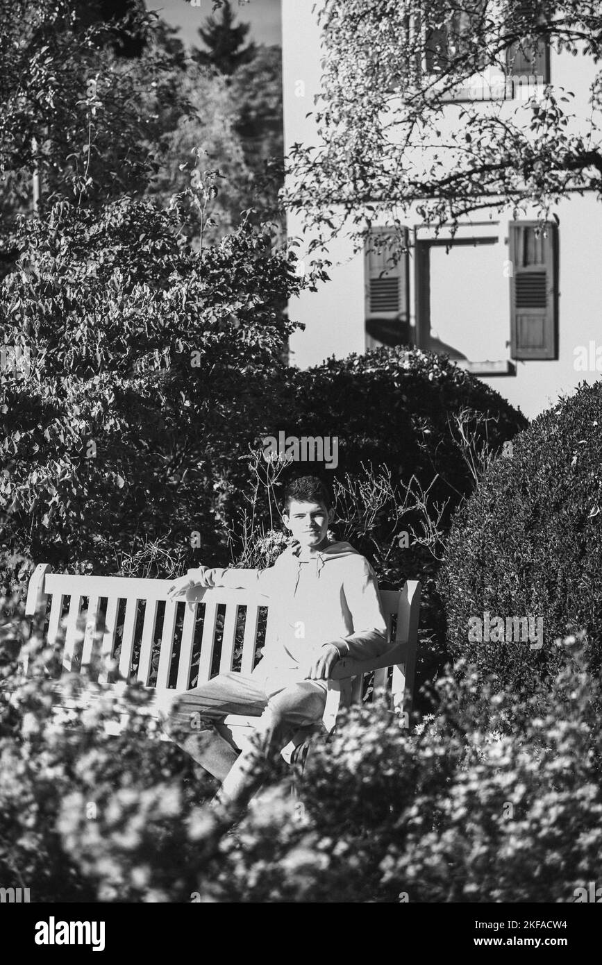 Un adolescente siede su Un banco nel Parco Autunno beve caffè da Un Thermo Mug e guarda in Un telefono. Ritratto di bel ragazzo allegro seduto Foto Stock