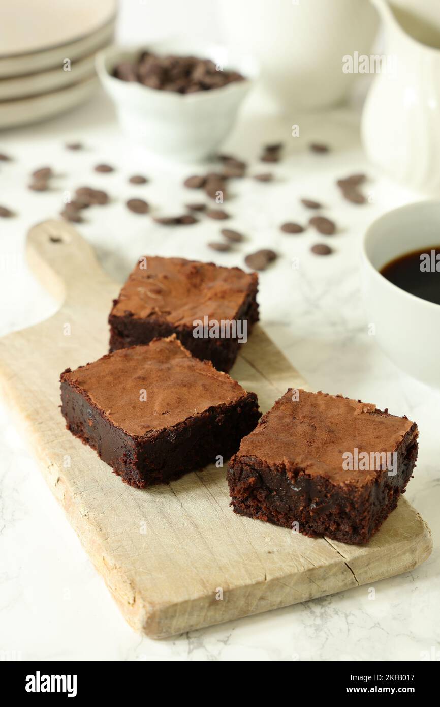 Brownie al cioccolato su un pannello di legno d'annata su sfondo bianco con tazza da caffè, piatti e gocce di cioccolato sparse Foto Stock