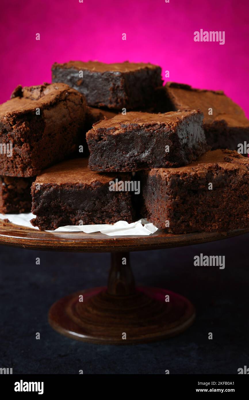 Una pila di brownie al cioccolato cotte in casa su una torta di legno vintage si contrappone a uno sfondo rosa intenso e vivace, ideale per San Valentino Foto Stock