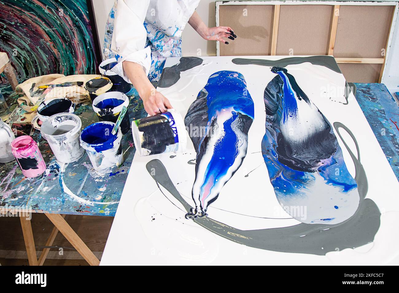 Foto ritagliata di mani di donna in vestito coperto di macchie versando vernice da secchio su tela con immagine astratta. Foto Stock