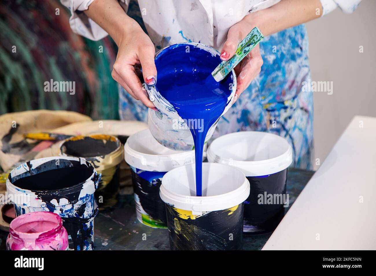 Foto ritagliata delle mani della donna in vestito coperto di macchie versando vernice acrilica blu dal secchio in un altro secchio. Foto Stock
