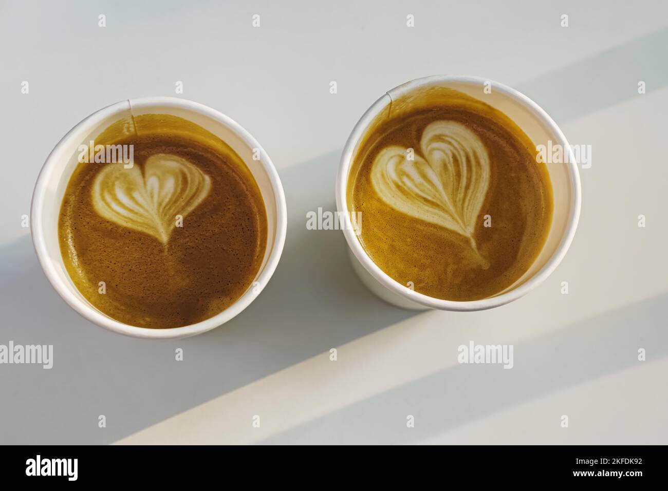 Due tazzine di carta di caffè con cuori disegnati sul tavolo bianco, vista dall'alto. Luce naturale del sole Foto Stock