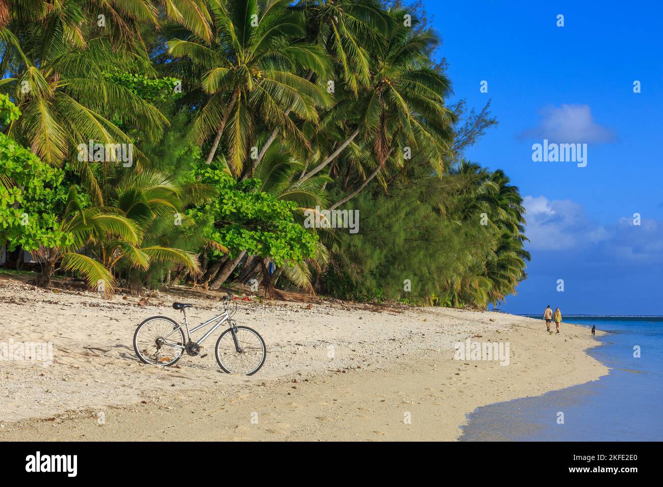 Una spiaggia tropicale, con una bicicletta parcheggiata sulla sabbia e una coppia passeggiando con un cane. Rarotonga, Isole Cook Foto Stock