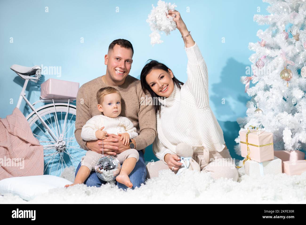 Una famiglia di tre persone si siede insieme nella zona foto di natale con decorazioni bianche su sfondo blu. Madre felice, padre e figlio piccolo hanno divertimento Foto Stock
