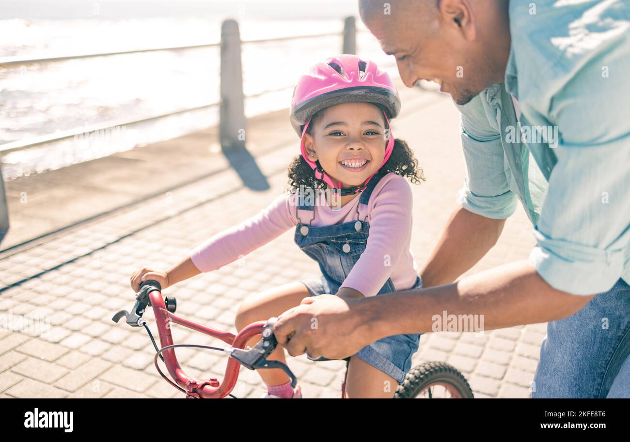 Ragazza nera, imparare e andare in bicicletta con papà sul lungomare con sorriso, casco o felice al sole. Bambino, padre e bicicletta in formazione, sicurezza o. Foto Stock