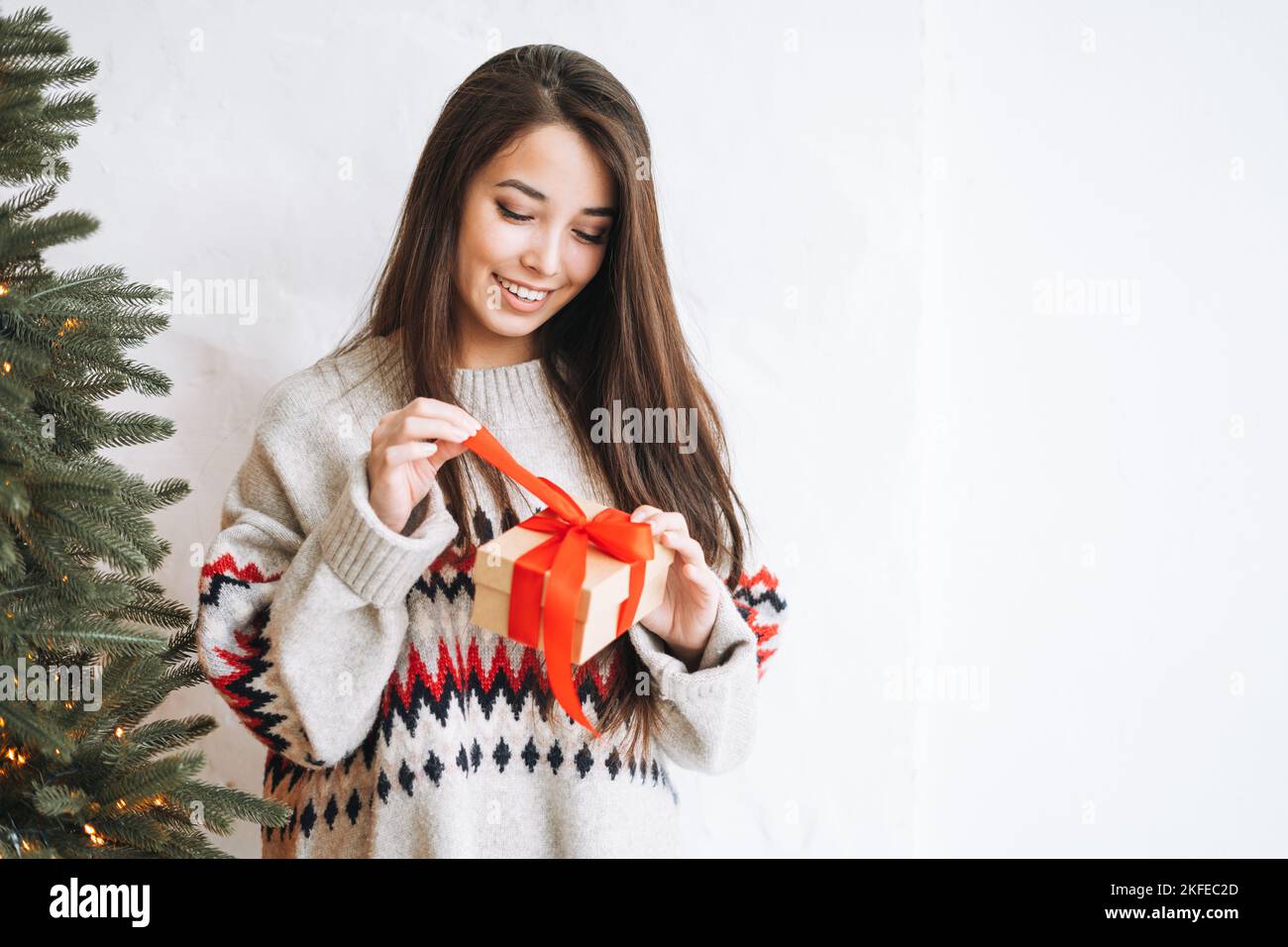 Giovane donna con capelli lunghi scuri in maglione accogliente con regalo scatola con nastro rosso in camera con albero di Natale a casa Foto Stock