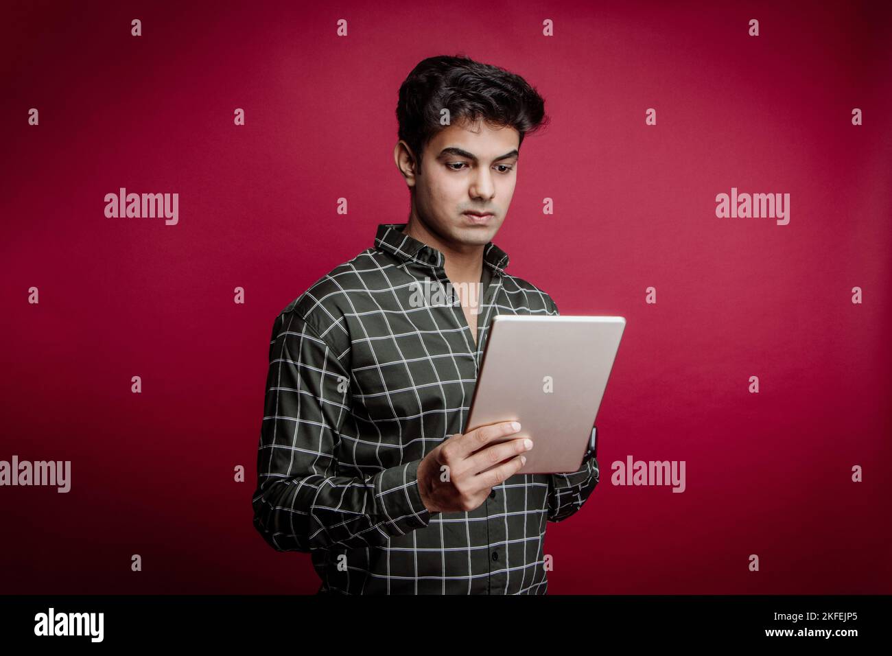 Un uomo indiano sicuro in una camicia a scacchi usando il tablet digitale contro la priorità bassa rossa che partecipa ad una riunione di affari in linea con i suoi clienti stranieri. Foto Stock