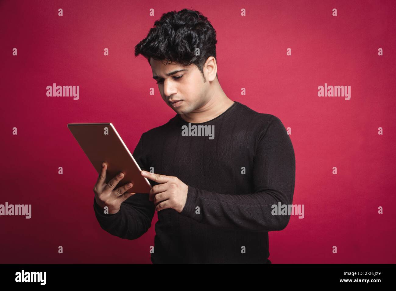 Serio giovane indiano in una t-shirt nera con un tablet digitale su sfondo rosso Foto Stock