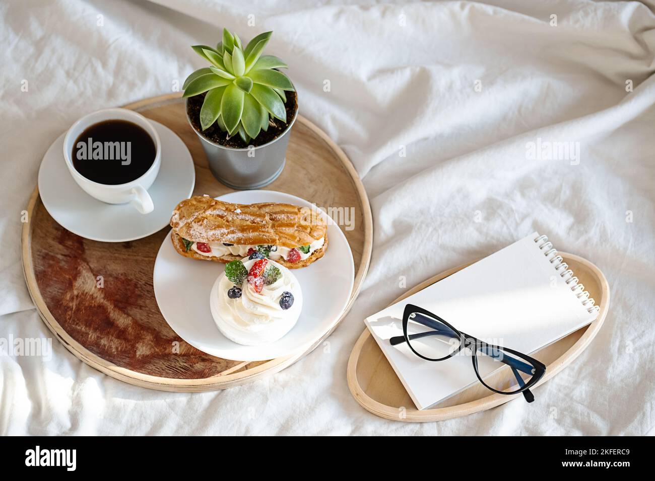 Torte, tazza da caffè, notebook e occhiali su vassoio di legno sulla biancheria da letto. Colazione accogliente a letto. Foto Stock