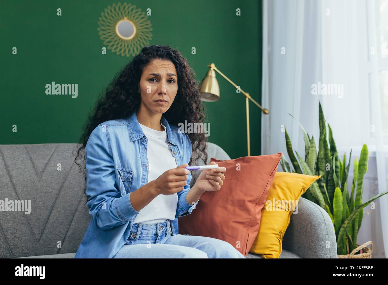 Una bella giovane donna latino-americana è sconvolta dalla notizia di una gravidanza non pianificata. È seduta a casa sul divano, tenendo un test di gravidanza a casa positiva nelle sue mani. Confuso, guarda la telecamera. Foto Stock
