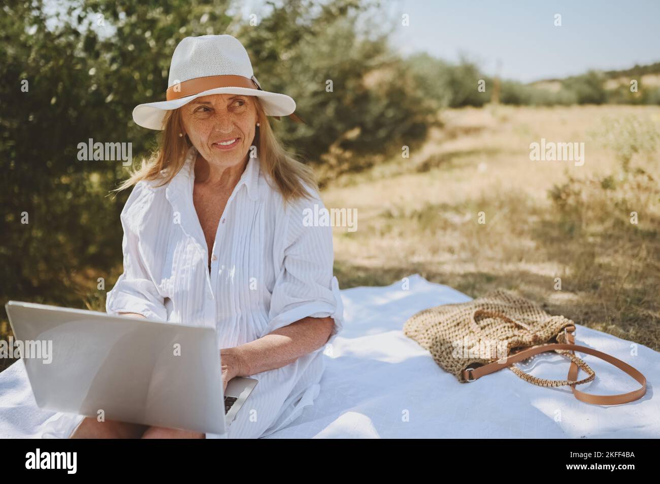 Donna anziana felice che lavora o videochiama con un computer portatile in estate all'aperto. Vecchia ed elegante signora in cappello di paglia e borsa seduta sull'erba durante il picnic in campagna. Concetto attivo delle persone in pensione Foto Stock