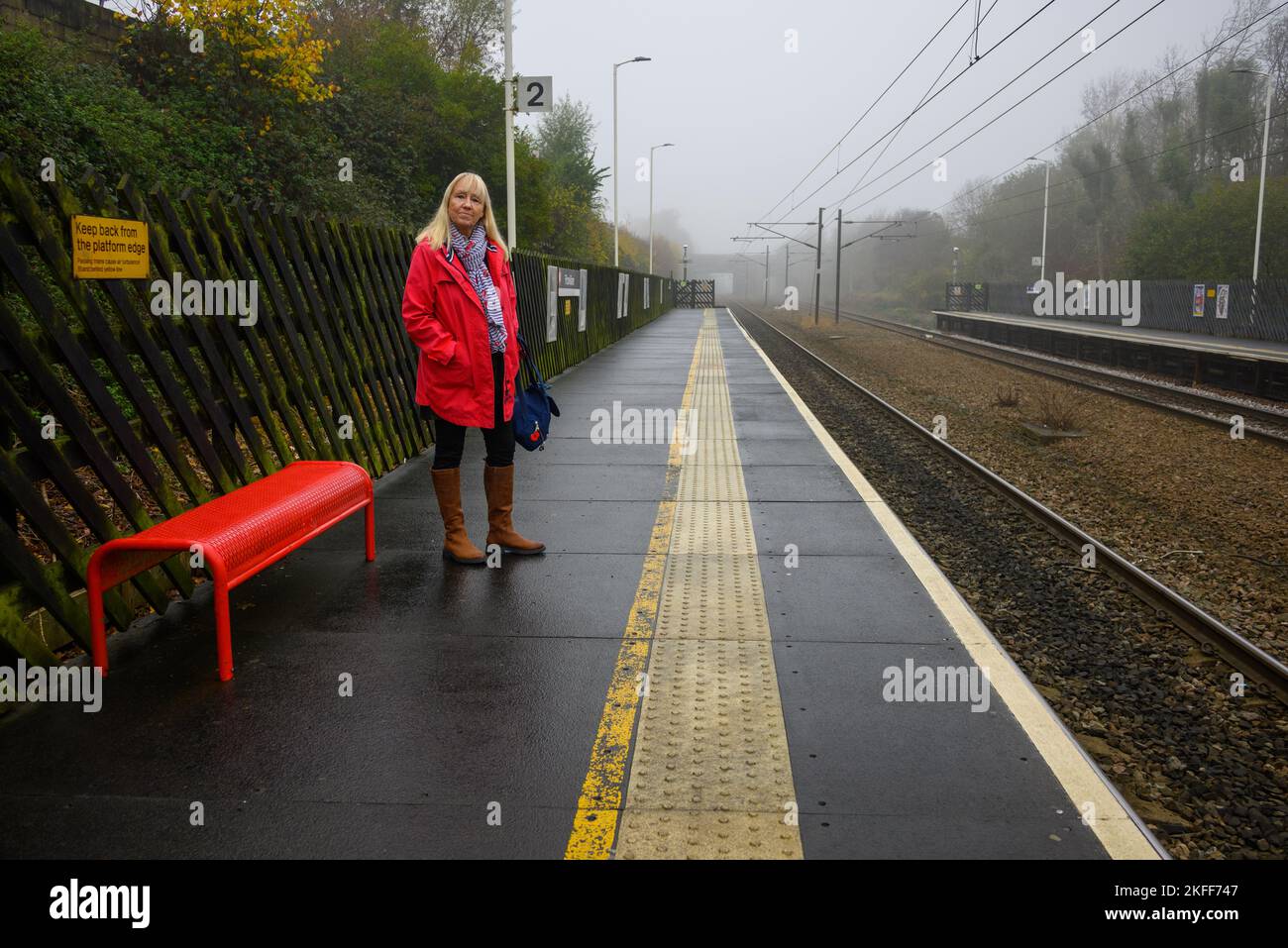 Donna in un cappotto rosso in piedi su una piattaforma nebbiosa nel mese di novembre in attesa di un treno Foto Stock