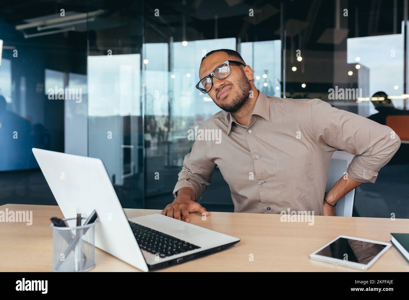 Uomo d'affari mal di schiena, uomo afroamericano massaggiante uomo di schiena che lavora seduto su sedia utilizzando un computer portatile al lavoro, all'interno di un moderno edificio per uffici. Foto Stock