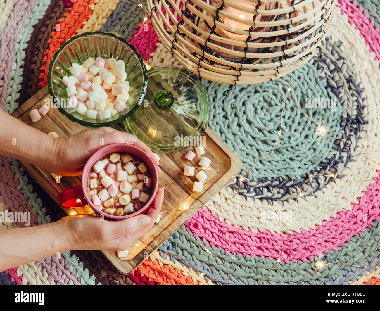 Vista ravvicinata delle mani delle donne che tengono una tazza rossa di cacao con mini marshmallows in un'accogliente giornata invernale, lanterna e luci a LED accese. Copia spazio su h Foto Stock