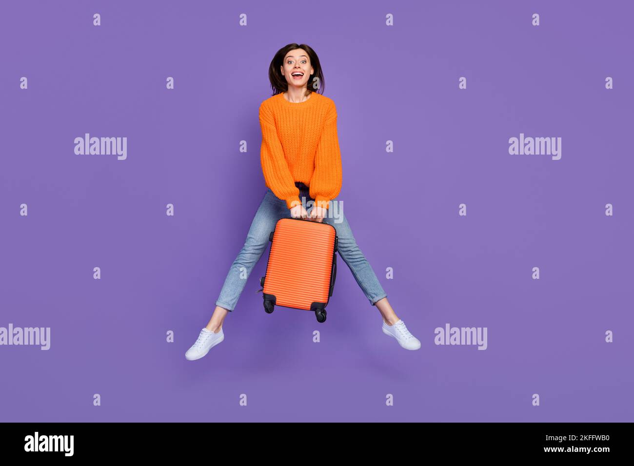 Foto a tutta lunghezza di carino giovane donna che salta bagaglio da viaggio vestita elegante abito a maglia arancione isolato su sfondo di colore viola Foto Stock
