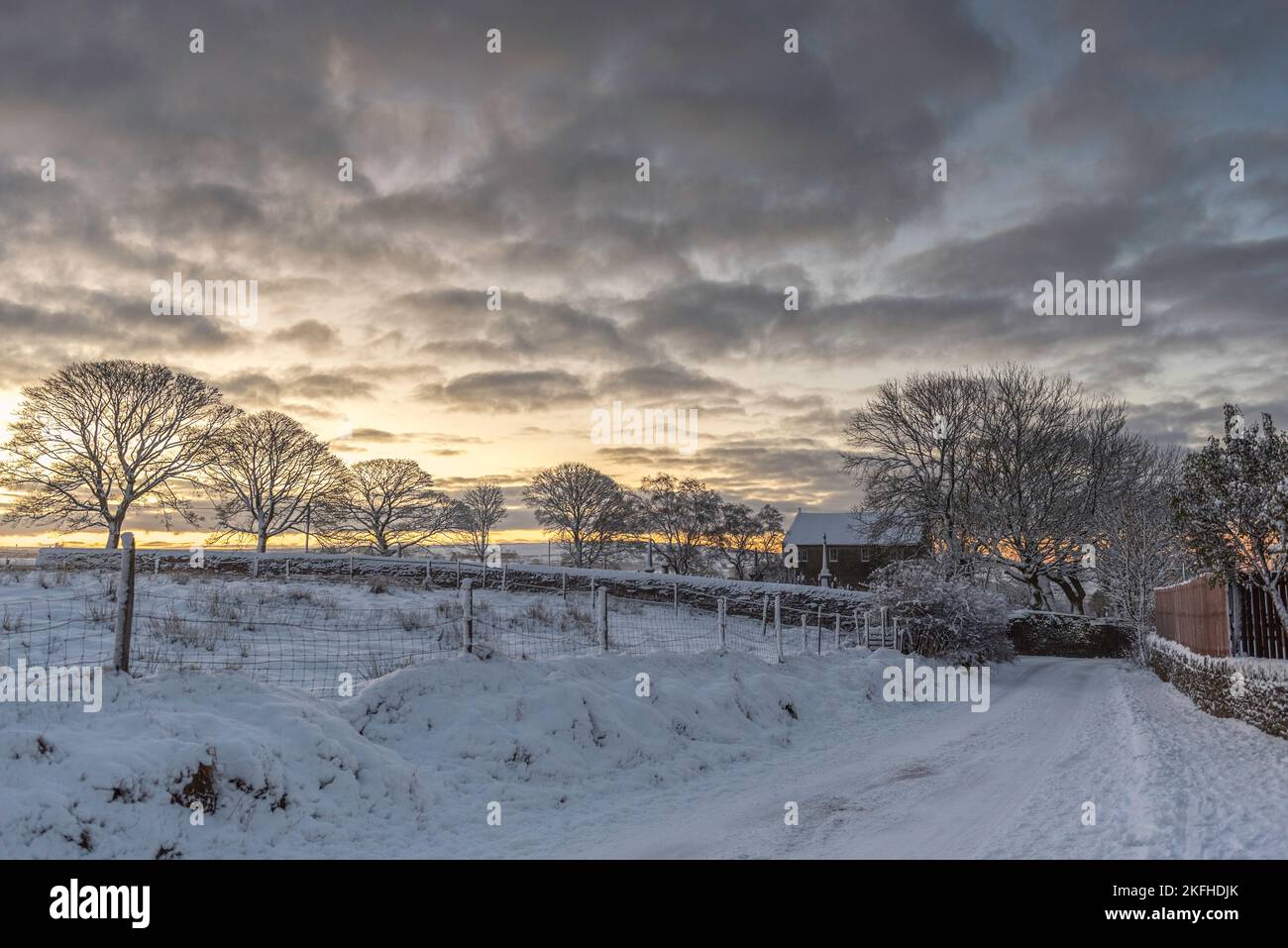 Vecchia corsia di campagna inglese nella neve d'inverno. Chiesa Battista di San Giovanni nella scena con le linee degli alberi e le nuvole piene di neve. Foto Stock