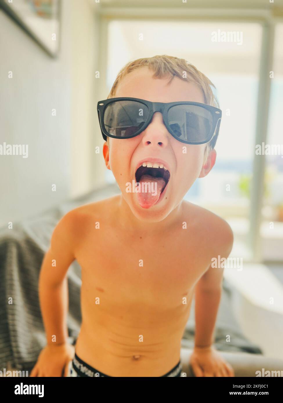 Ritratto di un ragazzo spensierato che indossa occhiali da sole neri che si stacca dalla lingua mentre si è in piedi in camera durante il fine settimana Foto Stock