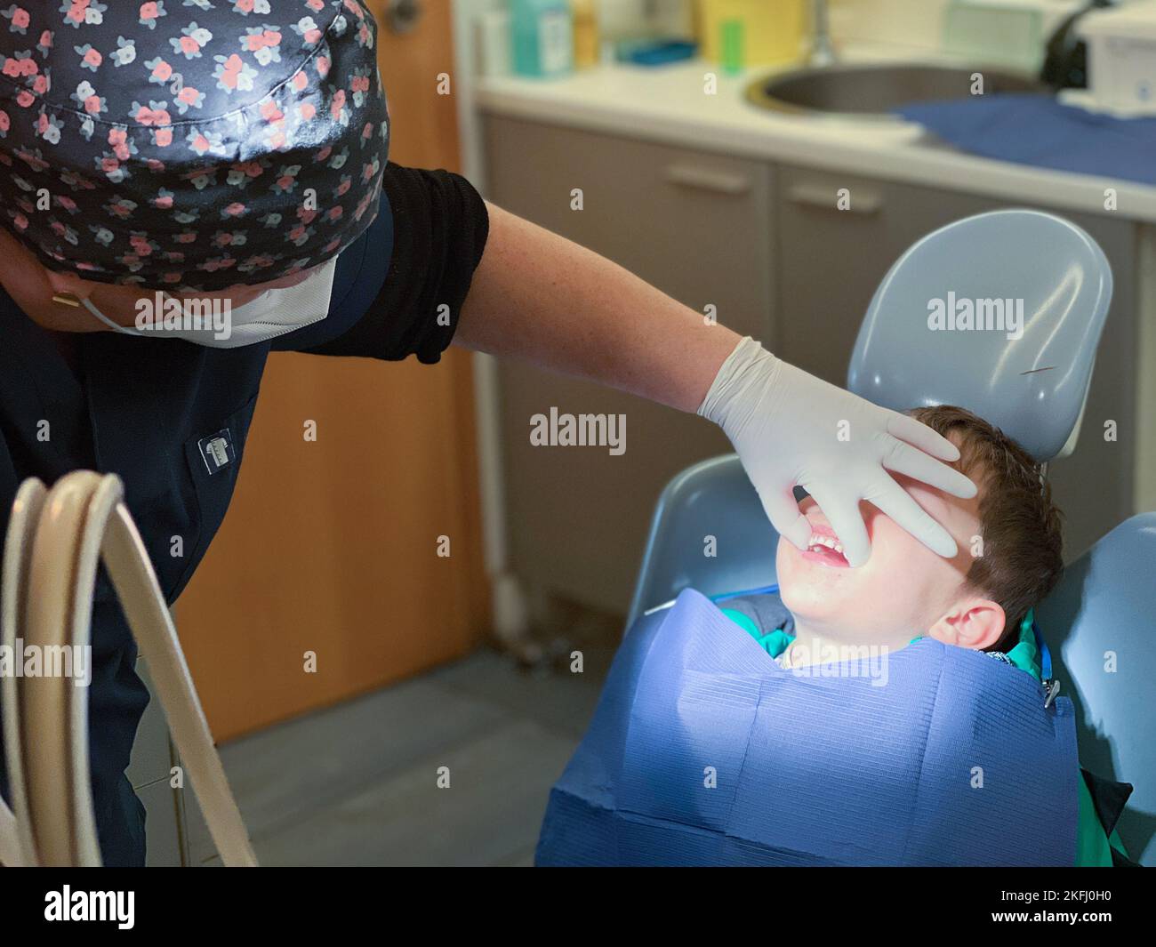 Dentista femmina che indossa un cappuccio floreale che tiene la bocca del ragazzo caucasico con la mano mentre esamina all'ospedale dentale Foto Stock