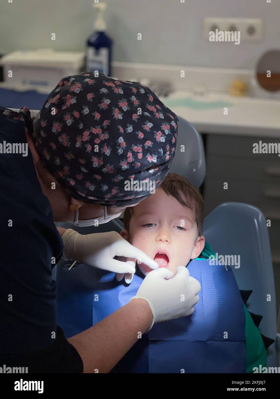 Dentista femmina che indossa cappuccio floreale e guanti chirurgici che esaminano i denti del ragazzo seduto sulla sedia in clinica dentale Foto Stock
