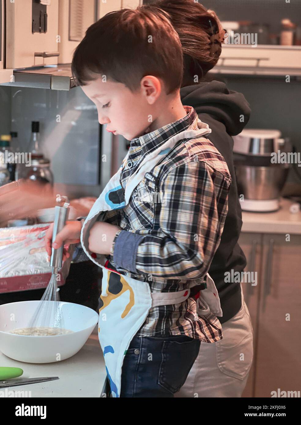 Vista laterale del simpatico ragazzo caucasico che indossa la pastella di miscelazione grembiule nel recipiente mentre si cuoce con la madre in cucina Foto Stock