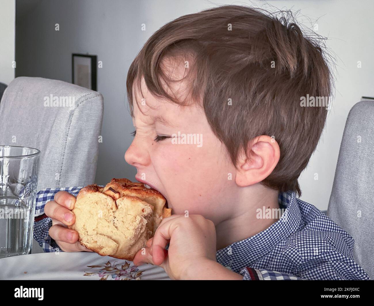 Vista laterale del ragazzo caucasico con gli occhi chiusi a prendere un boccone di pane mentre si faceva colazione sul tavolo da pranzo Foto Stock