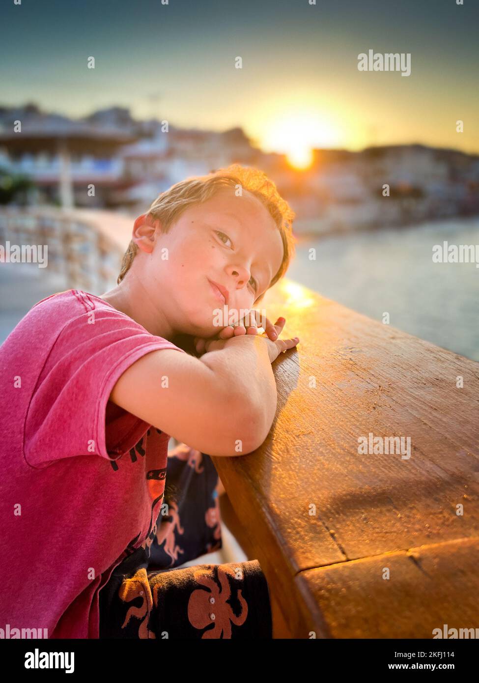 Ritratto del carino ragazzo caucasico che riposa la testa su una ringhiera di legno contro il bel cielo e il sole durante il tramonto Foto Stock