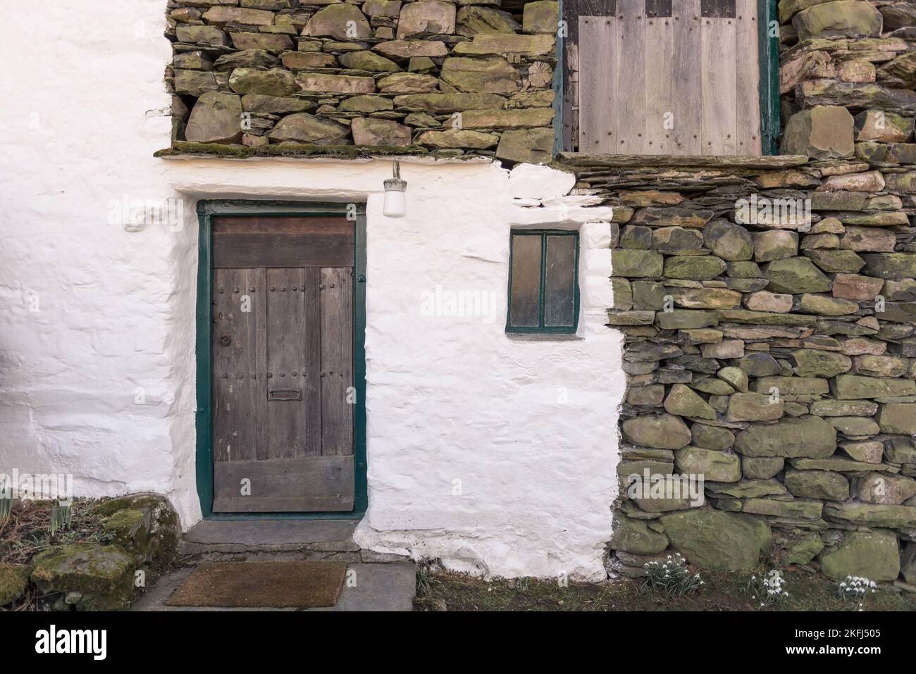 Vecchio cottage con pareti in pietra dipinta di bianco. Vecchia porta rustica in legno con fiori di neve a terra. Vecchia lampada rustica sopra la porta. Foto Stock