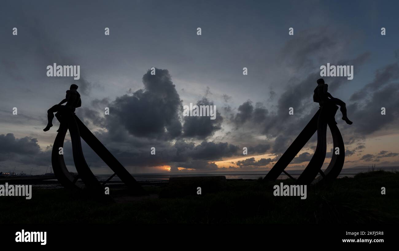Incredibile tramonto con splendide nuvole grigie nel cielo. Scultura a mezza luna a Heysham con uomini di metallo seduti sulla struttura che si affaccia sul mare. Foto Stock