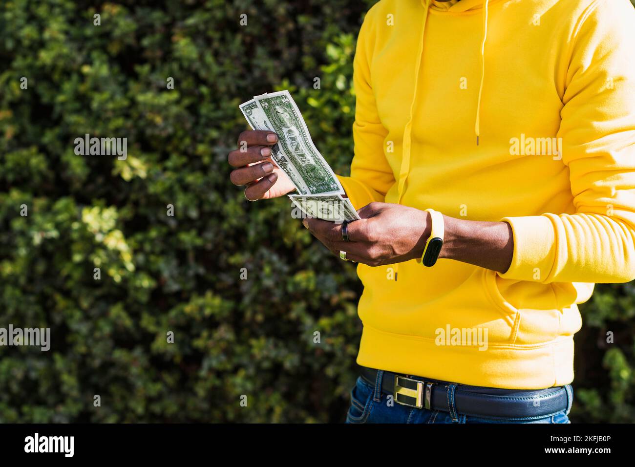 Primo piano delle mani di un uomo africano vestito con una felpa gialla e un braccialetto di attività irriconoscibile che tiene banconote in dollari mentre le passa davanti Foto Stock