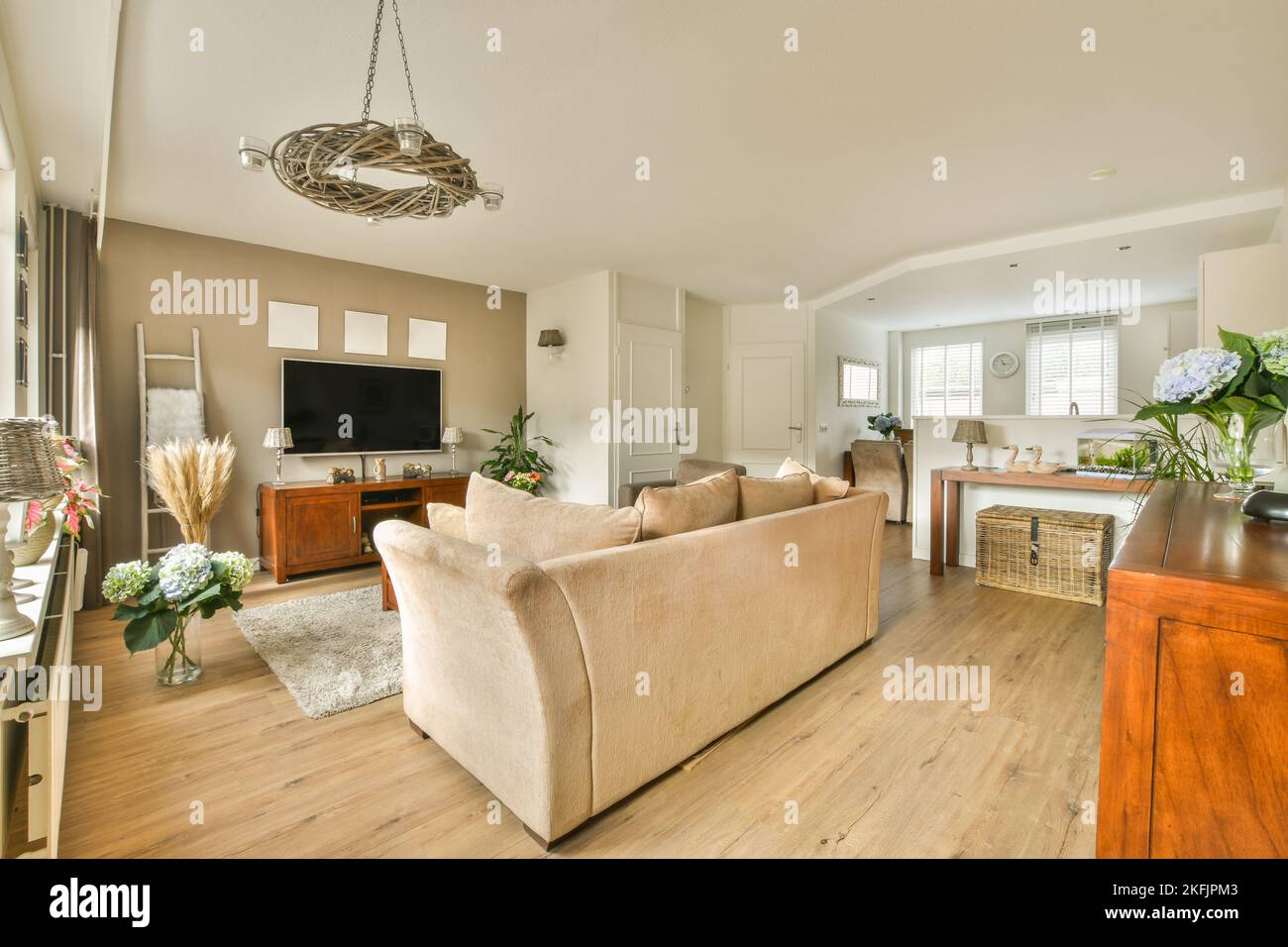 Divano ad angolo con morbidi cuscini situato sotto un elegante lampadario vicino alla TV nel luminoso soggiorno vicino alla cucina Foto Stock