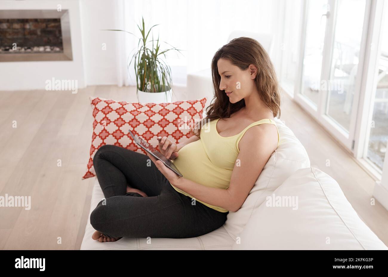Una giovane donna incinta che usa il suo tablet digitale mentre si siede sul divano del soggiorno. Foto Stock