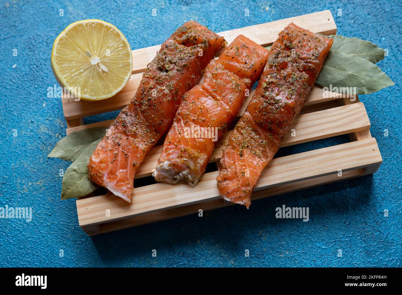 Tre pezzi di trota giacciono su una tavola su uno sfondo blu. Pesce cosparso di spezie. Nelle vicinanze si trova mezzo limone e una foglia di alloro. Foto Stock