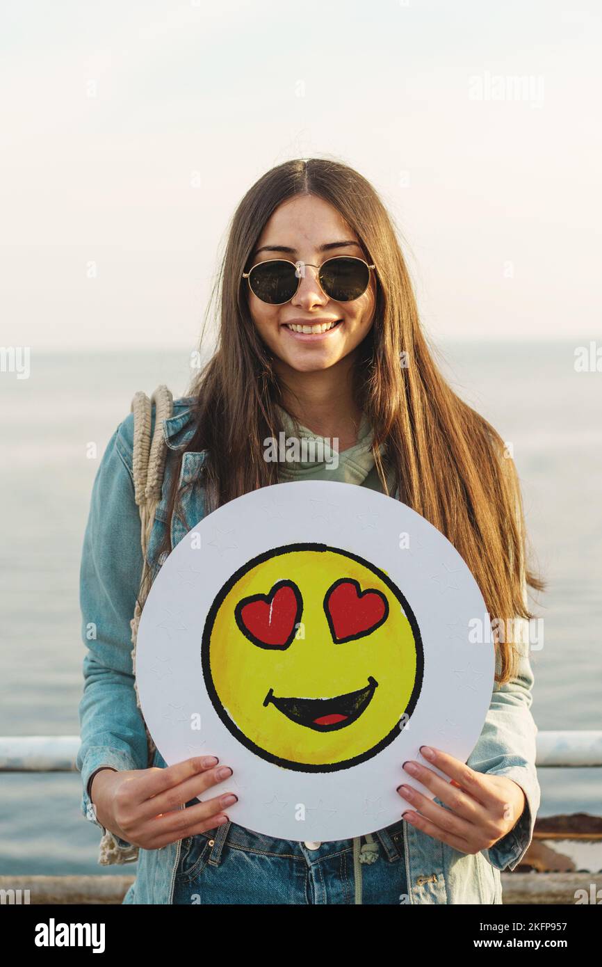 bella donna con occhiali da sole che tiene un emotoji icone sorridente all'aperto - persone e social network lifestyle concetto Foto Stock