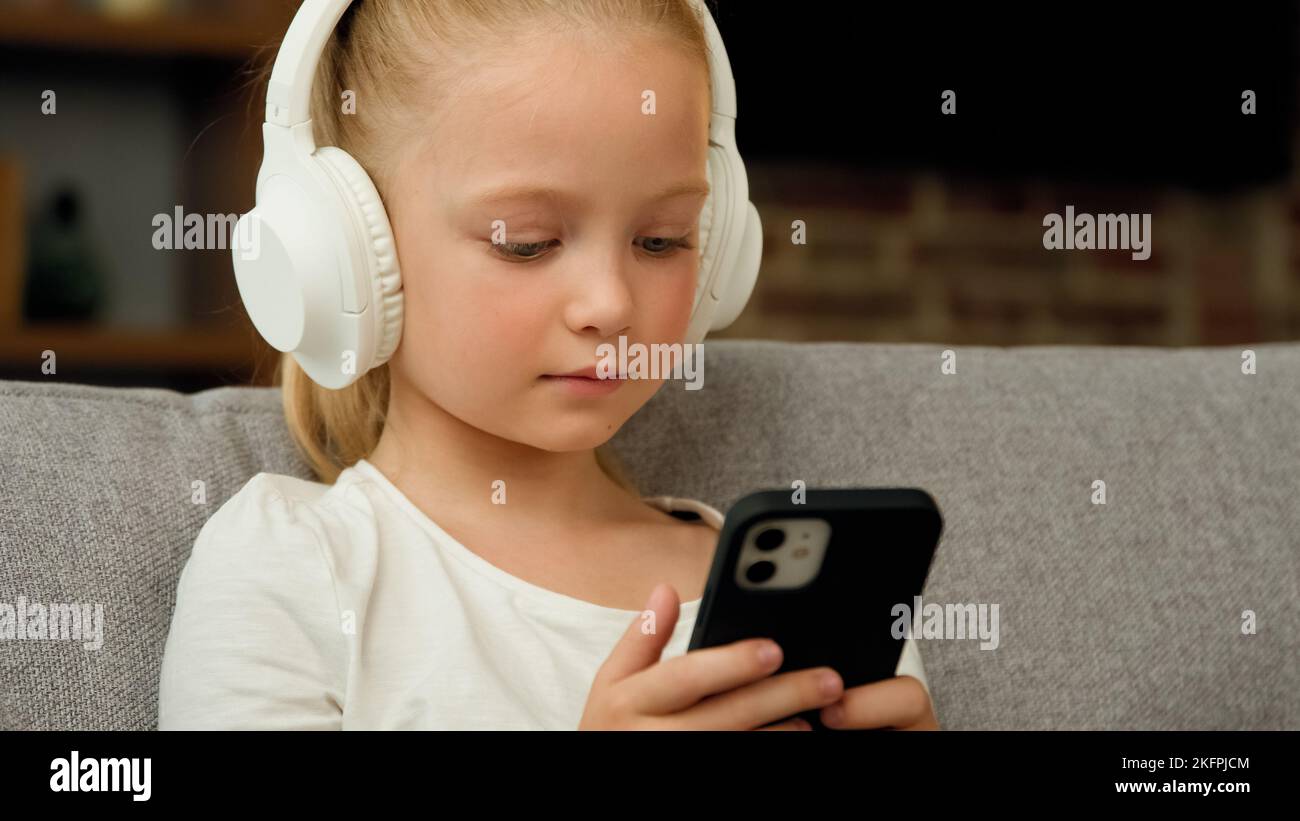 Cute ragazza caucasica bambino adolescente bambino sedersi sul divano rilassante ascoltare musica in cuffie guardare lo schermo del telefono cellulare godere la canzone pop preferita in audio Foto Stock