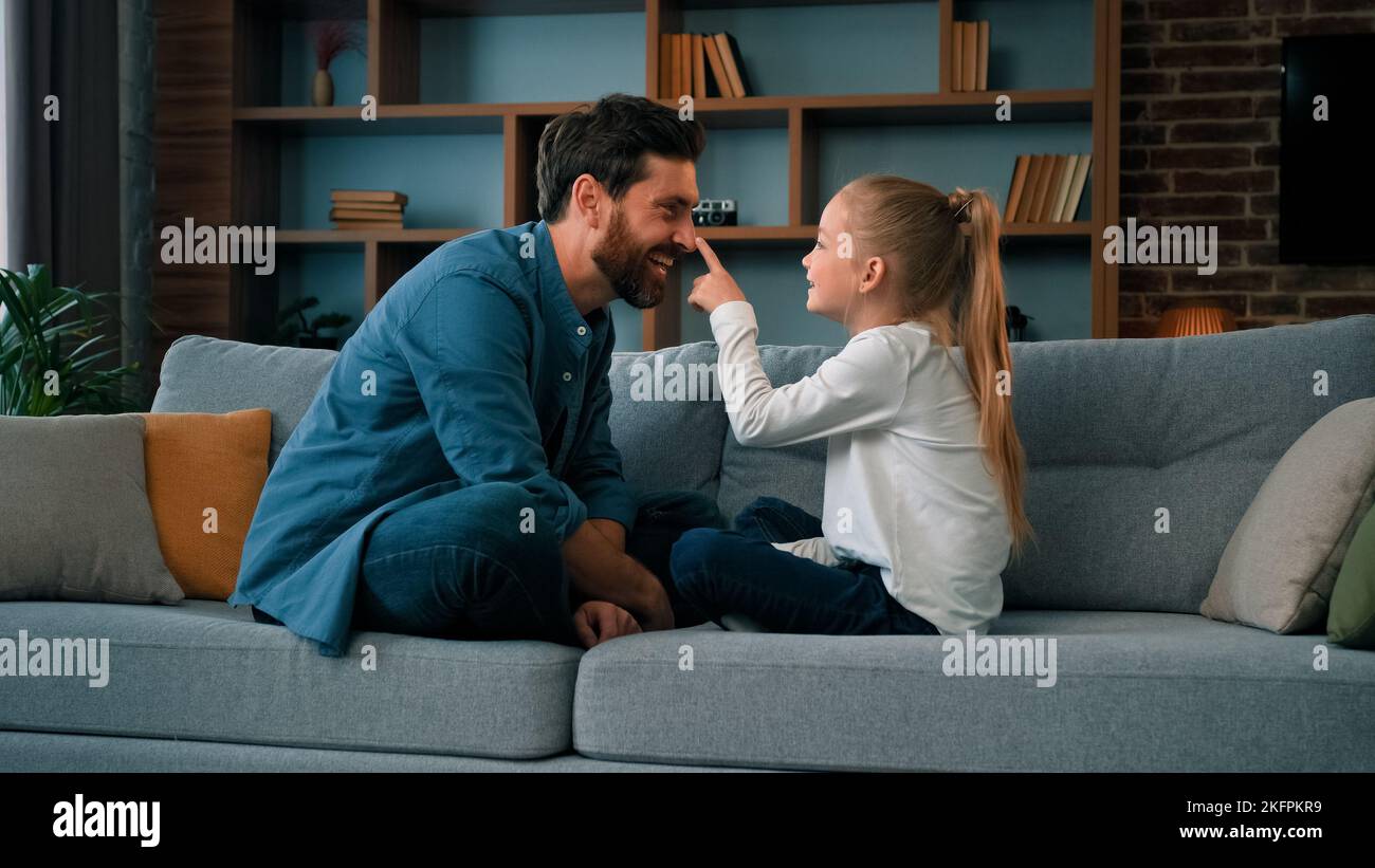 Divertente caucasico giovane famiglia papà e piccola figlia seduta confortevole sul divano amando ragazza carina toccare padre naso divertente scherzo giocare gioco Foto Stock