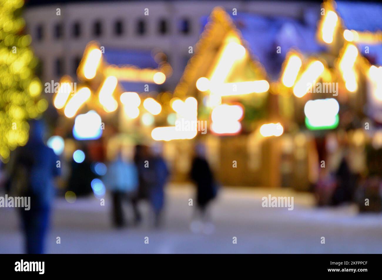 Sfondo sfocato. La gente cammina nella piazza della città la sera d'inverno. Silhouette nere di persone che camminano vicino a case decorate illuminazione luminosa. Luce bianca bokeh sfocare i punti dalle luci della casa incandescente Foto Stock